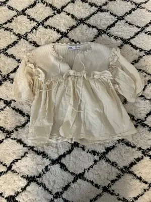 Supersöt beige blus från Zara med stickad överdel och volangdetaljer i spets längs axlarna. Blusen har puffiga korta ärmar och knytband framtill. Materialmix med stickad bomull upptill och luftig tyg nedtill. Perfekt för en romantisk look.