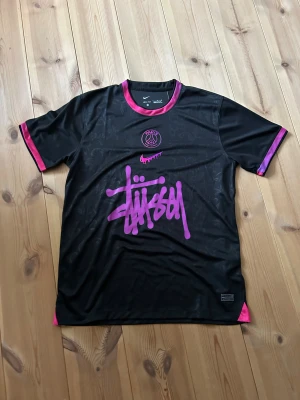 PSG x Stüssy svart fotbollströja Nike - Svart fotbollströja från Nike i samarbete med PSG och Stüssy. Tröjan har korta ärmar, rosa detaljer på krage och ärmslut, samt PSG-logga och Stüssy-grafik i rosa framtill. Materialet är lätt och andas med Dri-FIT-teknologi. Snyggt mönster i tyget för extra stil.