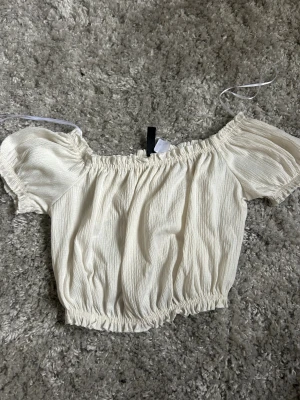 Vit offshoulder topp från H&M Divided - Supersöt vit offshoulder topp från H&M Divided i storlek XS. Toppen har puffiga korta ärmar och är gjord i ett lätt, krinklat bomullstyg med resår vid axlar och midja för en snygg passform. Perfekt för sommarens alla häng och selfies!