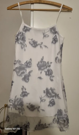 Vit blommig kortklänning med tunna axelband - Säljer en vit kortklänning med tunna axelband och gråa blommor som mönster. Klänningen har ett lätt och luftigt tyg med transparent nederkant, perfekt för dig som gillar romantisk och fräsch stil. Passar dig som vill ha något enkelt men ändå snyggt.