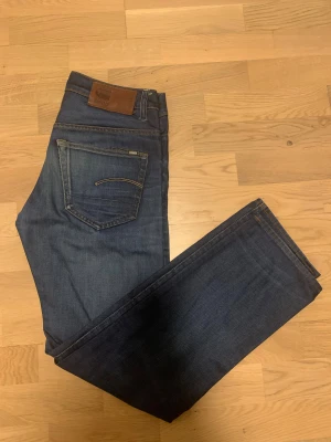Mörkblå jeans från G-Star RAW - Snygga mörkblå jeans från G-Star RAW med klassisk femficksdesign och bruna detaljer på bakfickan. Jeansen har raka ben, knappgylf och kontrastsömmar. Perfekt för dig som gillar stilrena och tidlösa jeans med coola detaljer. Det finns ett hål som syns tydligt på bilden , lätt att fixa den 