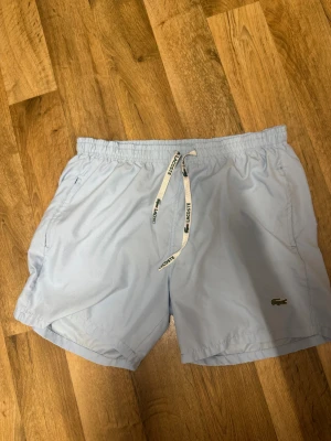Ljusblå badshorts från Lacoste - Snygga ljusblå badshorts från Lacoste med klassisk krokodillogga på benet och vit snörning med Lacoste-tryck i midjan. Shortsen har en loose passform och är perfekta för sommardagar vid stranden eller poolen.