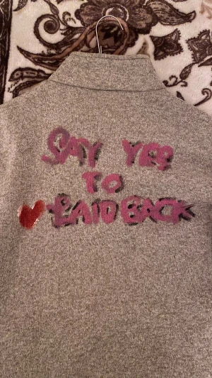 SAY YES TO LAIDBACK ( handgjord) Grå fleecejacka On The Peak strl 48 - Grå fleecejacka från On The Peak med svart dragkedja och detaljer i rosa på fickorna. Baksidan har graffiti-inspirerad text och ett rött hjärta. SAY YES TO LAIDBACK! Jackan har hög krage och praktiska fickor med dragkedja, perfekt för chill dagar.