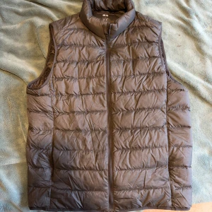 Grå dunväst från Uniqlo - Snygg brun dunväst från Uniqlo med quiltad design och hög krage. Västen har dragkedja framtill och är lätt och smidig att bära. Perfekt för lager-på-lager och håller dig varm under kyliga dagar. Enkel och stilren look som funkar till många outfits. Är skrynklig eftersom den har legat i min garderob ett tag men aldrig använd.