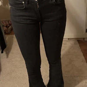 Svarta bootcut jeans - Säljer ett par svarta jeans med bootcut passform och låg midja. Klassisk femficksmodell med dragkedja och knapp framtill. Jeansen är tillverkade i stretchigt denim och har en snygg, mörk ton. Perfekt för dig som gillar en tidlös look. De är slitna vid benslutet då jag gått på dom men inget mer än de.
