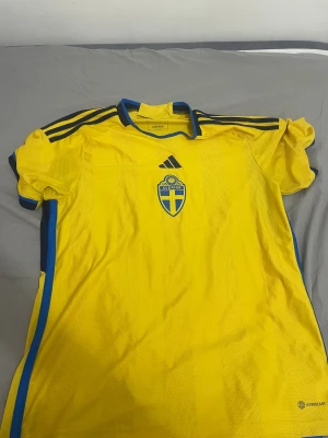 Svenska landslagets gul Adidas tröja L - Gul fotbollströja från Adidas i slim fit, officiell landslagströja för Sverige med blå detaljer och broderat landslagsemblem på bröstet. Tillverkad i lätt och ventilerande Aeroready-material, kortärmad och med klassiska Adidas-ränder på axlarna.
