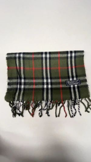 Rutig grön halsduk från Burberrys - Snygg rutig halsduk från Burberrys i klassisk grön, svart, vit och röd färg. Gjord av 100% lammull och har fransar i ändarna. Perfekt accessoar för att lyfta din stil med ett brittiskt touch.