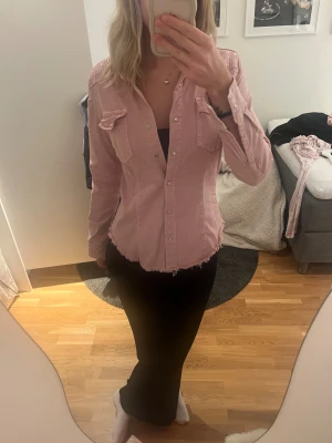 Rosa jeansjacka med paljettdetaljer - Rosa jeansjacka med långa ärmar, knappar framtill och två bröstfickor. Skjortan har paljettdetaljer på axlar och rygg samt rå, fransig nederkant för en cool look. Figurnära passform och klassisk krage. Perfekt för dig som gillar unika detaljer.