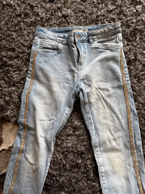 Skinny ljusblå jeans med glitterdetalj - Snygga och tuffa ljusblå skinny jeans med guldglittrande ränder längs båda benen. Klassisk femficksmodell med knapp och dragkedja framtill. Jeansen har en smal passform och är tillverkade i mjukt denimtyg som sitter skönt hela dagen.