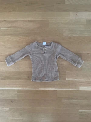 Brun ribbad långärmad tröja H&M - Säljer en brun långärmad tröja från H&M i ribbad bomull. Tröjan har två knappar vid halsen och ett snyggt, grovt stickat mönster som ger en chill vibe. Perfekt för lager-på-lager under hösten och vintern.