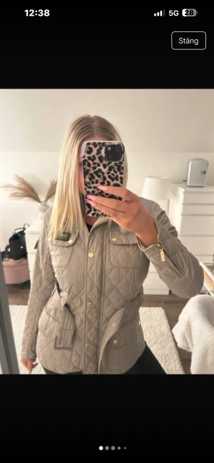 Beige quiltad jacka från Barbour - Snygg beige quiltad jacka från Barbour med bälte i midjan och guldfärgade knappar. Jackan har lång ärm, krage och praktiska fickor framtill. Perfekt för dig som gillar klassisk stil med modern touch.