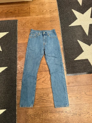 Levi's 501 blå jeans W25 L30 - Klassiska Levi's 501 jeans i ljusblå tvätt med raka ben och fem fickor. Modellen har hög midja och ikonisk röd Levi's-tag på bakfickan samt läderpatch. Jeansen är tillverkade i slitstarkt bomullsjeansmaterial och har knappgylf framtill. tvättade max 2 gånger.