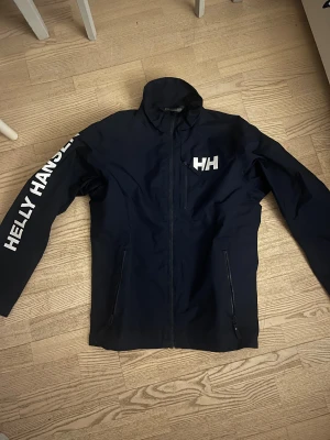 Mörkblå vindjacka från Helly Hansen - Snygg mörkblå vindjacka från Helly Hansen med vit logga på bröstet och ryggen samt stort tryck på ena ärmen. Storleken är S, använd en tid Men inte i dåligt skick, skulle säga att konditionen är 8/10. 