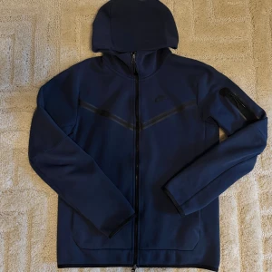 Nike tech fleece  - Mörkblå hoodie med zip från Nike med hel dragkedja och huva. Jackan har svarta detaljer över bröstet och en ficka med dragkedja på ärmen. Tillverkad i bomull och polyester för en mjuk och sportig känsla.