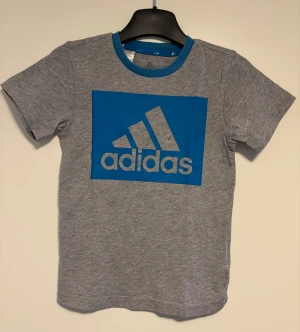Adidas t-shirt strl 98 - Snygg grå t-shirt från Adidas med stor blå logga framtill. Rund hals med blå ribbad kant och klassisk kortärmad passform. 