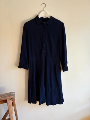 Marinblå skjortklänning Tommy Hilfiger - Snygg marinblå skjortklänning från Tommy Hilfiger i mjuk viskos. Klänningen har klassisk krage, knappar framtill och långa ärmar med manschett. En knapp som ska sitta högst upp saknas, se bild. 