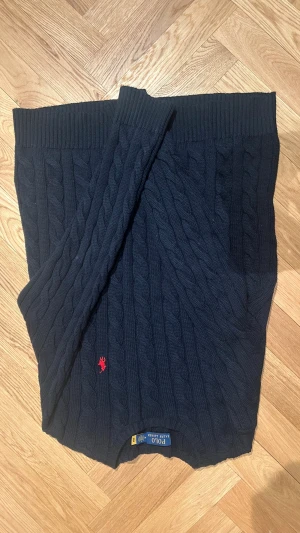 Mörkblå stickad tröja från Polo Ralph Lauren - Säljer en mörkblå stickad tröja från Polo Ralph Lauren med klassiskt kabelstickat mönster och röd broderad logga på bröstet. Tröjan har ribbade muddar och rund hals, perfekt för dig som gillar stilren och tidlös design.