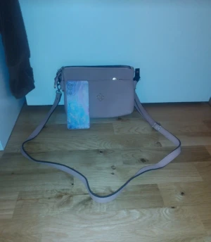 Ljusrosa axelväska med dragkedja - Snygg ljusrosa axelväska med justerbar rem och dragkedja upptill. Väskan har en stilren design med ett litet emblem på framsidan och är tillverkad i skinnimitation. Perfekt storlek för mobil och småsaker, passar dig som gillar enkel och clean look.