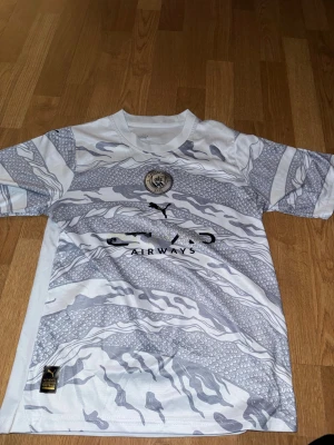 Manchester City x Puma vit matchtröja - Säljer en officiell Manchester City fotbollströja från Puma i vitt med grått mönster. Tröjan har korta ärmar, rund hals och coolt silverfärgat klubbmärke på bröstet. Materialet är lätt och ventilerande polyester, perfekt för träning eller match.