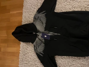 Svart hoodie Marcelo Burlon M - Svart hoodie från Marcelo Burlon County of Milan i storlek M. Tröjan har ett grått grafiskt mönster över axlar och bröst, huva med snörning och en stor magficka. Tillverkad i 100% bomull för skön och avslappnad känsla.