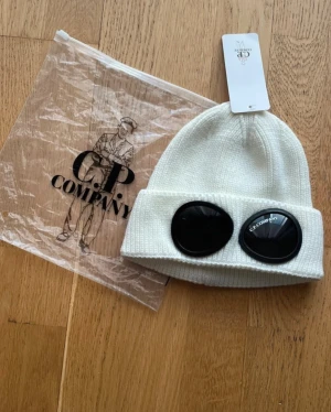 Cp  Company  mössa - Säljer en vit ribbstickad mössa från C.P. Company med ikoniska svarta goggle-linser på uppviket. Mössan har en clean och modern look, perfekt för dig som gillar streetwear och vill sticka ut. Materialet är mjukt och stretchigt för skön passform.