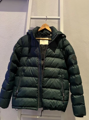 Mörkgrön pufferjacka från Gant - Snygg mörkgrön pufferjacka från Gant. Jackan har två sidofickor med dragkedja och diskret logga på bröstet samt ett märke på ärmen. Perfekt för kalla dagar. Priset är diskuterbart och skriv gärna för fler bilder.