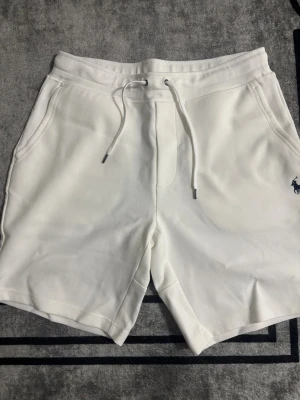 Vita shorts från Polo Ralph Lauren - Snygga vita shorts från Polo Ralph Lauren med klassisk broderad logga i marinblått på vänster ben. Modellen har snörning i midjan, två sidofickor och en bakficka. Mjukt material i bomull som är perfekt för chill dagar. Passformen är normal och midjan är midwaist.