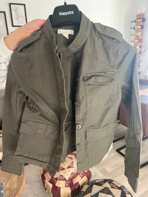 Olivgrön utilityjacka från H&M, stl 38 - Snygg olivgrön utilityjacka från H&M i storlek 38. Jackan har flera stora fickor framtill, axelklaffar och knäppning med knappar. Tillverkad i bomull och har en rak, lite loose passform. Perfekt för dig som gillar en avslappnad och cool stil.