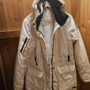 Beige parkajacka med huva och svarta detaljer - Snygg vit parkajacka med huva och svarta dragkedjor. Jackan har flera praktiska fickor. Perfekt för kyliga dagar och har en stilren look med kontrasterande svarta detaljer.