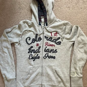 Grå hoodie med tryck från Miss America - Grå hoodie från Miss America med broderat och tryckt text på framsidan. Tröjan har dragkedja, snörning i huvan och fickor framtill. Insidan av huvan har ett mönstrat tyg i blått och rött. Perfekt för chill dagar och streetstyle.