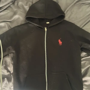 Svart hoodie från Polo Ralph Lauren M - Svart hoodie från Polo Ralph Lauren i storlek M. Tröjan har dragkedja framtill, huva och en röd broderad logga på bröstet. Tillverkad i mjuk bomullsmix med fleece på insidan för extra komfort. Perfekt för chill dagar eller när du vill ha en clean streetstyle. Ja vet inte om den är äkta eller inte för de är inte min tröja.