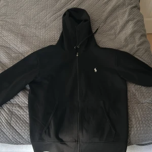 Svart hoodie från Polo Ralph Lauren - Svart hoodie från Polo Ralph Lauren med dragkedja och klassisk broderad logga på bröstet. Tröjan har huva med snörning och två fickor framtill. Perfekt för en chill och stilren look.