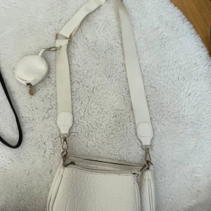 Vit väska med krokopräglat mönster - Säljer en vit väska med bred axelrem och krokopräglat mönster. Väskan har flera fack med dragkedja och en liten rund miniväska som sitter fast på remmen. Perfekt för dig som vill ha en trendig och praktisk väska med snygga detaljer.