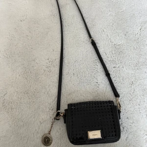 Svart vintage axelremlväska från DKNY i gländigskinn - Snygg svart vintage äkta axelväska från DKNY i glansigt skinn med rutigt perforerat mönster på locket. Väskan har justerbar axelrem, guldfärgade metalldetaljer och ett dekorativt DKNY-mynt. Invändigt finns flera kortfack och DKNY-logga på fodret.