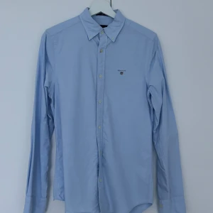 Ljusblå skjorta från GANT - Snygg ljusblå skjorta från GANT med klassisk button-down krage och broderad logga på bröstet. Skjortan är tillverkad i 100% bomull, sydd i Oxford stil och har långa ärmar samt knappar framtill. Perfekt för dig som gillar stilrena och tidlösa plagg.