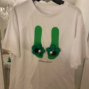 Vit t-shirt med 3D-blommor och pärlor - Unik vit t-shirt med grönt tryck framtill och två 3D-blommor i grönt och svart, dekorerade med stora pärlor. Texten 'A CHEMISTRY OF SELF NO' under motivet. Klassisk passform och korta ärmar. Perfekt för dig som vill sticka ut med coola detaljer.