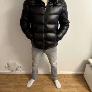 Moncler Maya - Säljer en svart pufferjacka från Moncler med huva och dragkedja. Jackan har Moncler-logga på ärmen, två sidofickor och quiltad design. Materialet är glansigt och fyllningen är dun, vilket gör den perfekt för kalla dagar. Klassisk och stilren look.