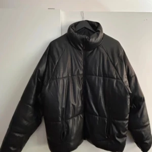 Svart pufferjacka från Bershka - Säljer en svart pufferjacka från Bershka i syntetmaterial med skinnlook. Jackan har hög krage, dragkedja framtill och två fickor med dragkedja. Perfekt för kalla dagar och ger en clean, modern vibe.