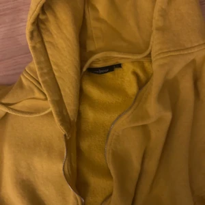 Gul hoodie med dragkedja från Jacoe - Gul hoodie från Jacoe med dragkedja framtill och huva med snörning. Tröjan har långa ärmar, ribbade muddar och resår i midjan. Perfekt för dig som gillar färg och vill ha en skön och avslappnad stil.