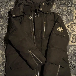 Svart dunjacka från Moose Knuckles - Säljer en svart dunjacka från Moose Knuckles med huva, silverfärgade dragkedjor och tryckknappar. Jackan har flera fickor, logga på ärmen och en schysst puffer-look. Perfekt för kalla dagar när du vill hålla stilen.