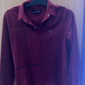 Vinröd pikétröja Emporio Armani S - Snygg vinröd långärmad pikétröja från Emporio Armani med klassisk krage och knappar framtill. Tröjan har diskreta svarta linjer som mönster och den ikoniska Armani-loggan på bröstet. Perfekt för dig som vill ha en stilren och exklusiv look.