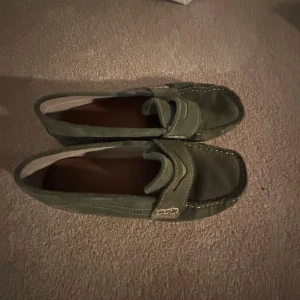 Gröna mockaloafers från Zara stl 33 - Snygga gröna loafers från Zara i mocka med klassisk design och kontrastsömmar. Skorna har platt sula och dekorativ detalj på ovandelen. Perfekta för dig som gillar stilrena och bekväma skor med en twist.