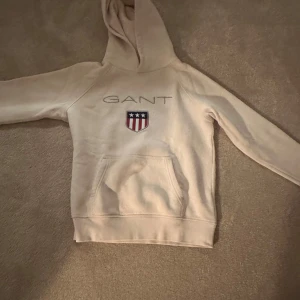 Beige hoodie från GANT med brodyr - Snygg beige hoodie från GANT med broderad logga och sköld på bröstet. Tröjan har huva, magficka och långa ärmar. Perfekt för dig som gillar klassisk och stilren streetwear.