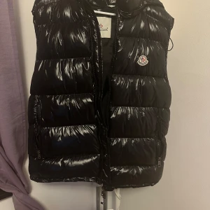 Svart dunväst med huva - Snygg svart dunväst från Moncler med glansig finish och avtagbar huva. Västen har dragkedja framtill, två fickor med dragkedja och klassisk Moncler-logga på bröstet. Perfekt för lager-på-lager och streetstyle.