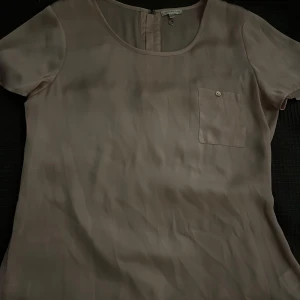 Ljusrosa blus från Stockh LM - Säljer en stilren ljusrosa blus från Stockh LM i storlek 40. Blusen har korta ärmar, rund halsringning och en bröstficka med knappdetalj. Materialet är slätt och känns lyxigt, och den har dragkedja i ryggen för enkel påklädning.