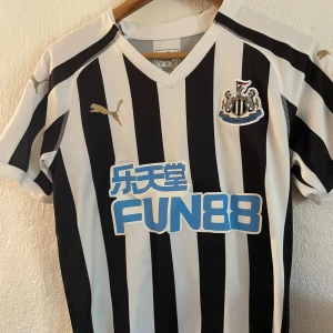 Newcastle United randig fotbollströja Puma - Snygg Newcastle United fotbollströja från Puma med klassiska svarta och vita ränder. Tröjan har klubbmärke på bröstet, guldiga Puma-loggor och sponsortryck i blått. Tillverkad i lätt och ventilerande material, perfekt för match eller träning.