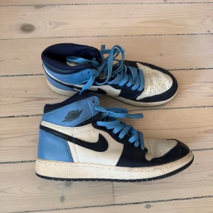 Nike Air Jordan 1 Obsidian - Säljer ett par Nike Air Jordan 1 sneakers i blått, vitt och marinblått skinn. Klassisk high-top modell med blå snörning och svart swoosh. Perfekta för dig som gillar streetwear och vill sticka ut med en ikonisk siluett. Gott skick, köpt i Kanada, kan skickas eller hämtas :)