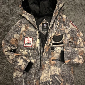 Canada Goose x OVO camo dunjacka - Säljer en Canada Goose x October's Very Own dunjacka med camouflagemönster i bruna och grå toner. Jackan har huva, flera fickor, patchar på bröst och ärm samt dragkedja och knappar. Perfekt för kalla dagar och riktigt unik stil. Passar storlek XS-S