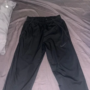  Nike Dri-FIT Woven byxor - Svarta träningsbyxor från Nike med Dri-FIT-teknologi och standard fit. Byxorna har elastisk midja, sidofickor och en diskret Nike-logga på benet. Perfekta för dig som vill ha bekväma och snygga byxor till träning eller chill.