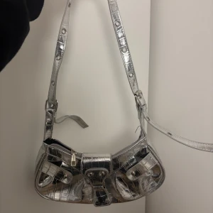 Silvrig axelväska i croco-mönster - Snygg axelväska i metallic-silver med croco-mönstrad yta och justerbar rem. Väskan har flera fickor framtill med spännen och dragkedja upptill. Perfekt för dig som vill sticka ut med en trendig accessoar.